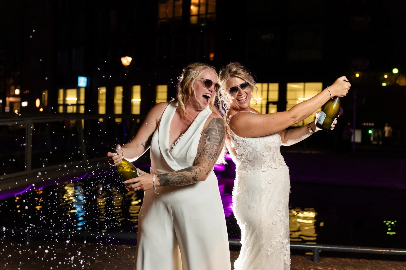 Twee bruiden spuiten lachend champagne in de avond bij de fontein